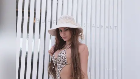 AndreaKalo wears a leopard print bikini and a white sun hat, posing against a white slatted wall with her long brown hair down.