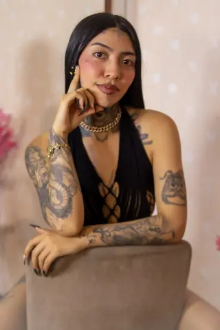 Tattooed woman in a black halter top posing thoughtfully in a soft-lit indoor setting.