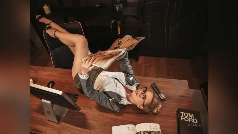 BeaNix lies on a wooden desk wearing a grey blazer and white shirt, holding an open magazine over her face.