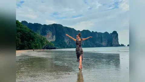 AnaMathers stands in a long, dark patterned dress on a wet sandy beach with her arms outstretched against a backdrop of large tropical cliffs.