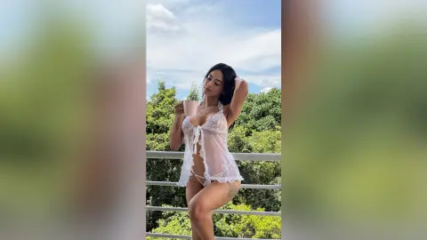 Young woman in white lace lingerie posing on a balcony overlooking lush green trees under a bright sky.