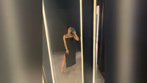 Stylish woman in a sleek black midi dress taking a mirror selfie in a modern, dimly lit room.