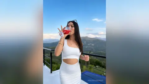 Young woman in a white summer outfit sipping a red cocktail on a scenic mountain balcony at sunset.
