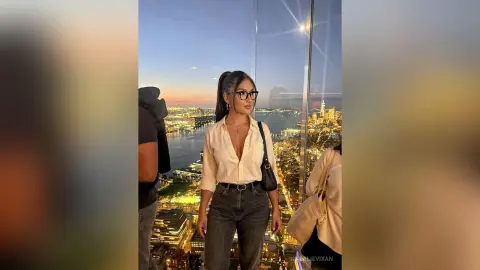 Stylish woman in a white blouse and jeans overlooking the glowing New York City skyline at sunset.