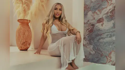 Blonde woman in a white sheer mesh outfit sitting elegantly in a bright, modern minimalist room.