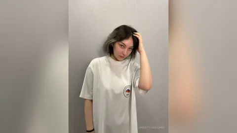 Young woman in a white oversized graphic t-shirt posing against a minimalist grey wall with a relaxed mood.