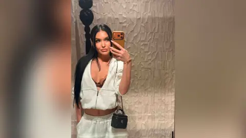 Young woman with long dark hair in a white lounge set taking a mirror selfie in a modern textured room.
