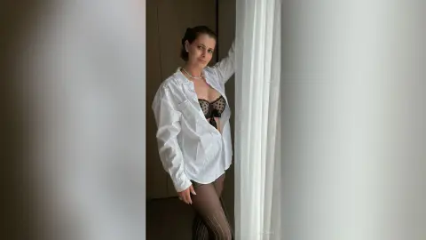 Smiling woman in a white button-down shirt and black lingerie posing by a window in a bright room.