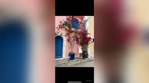 Woman in a white summer dress standing by a pink building with blooming bougainvillea in a sunny Mediterranean setting.