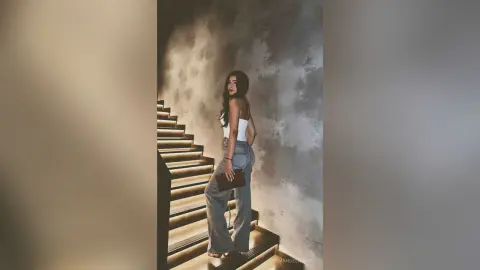Stylish woman in white tank top and baggy jeans holding a clutch on a moody, dimly lit staircase.