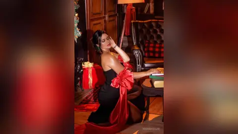 Elegant woman in a black evening gown with red accents posing in a cozy, festive living room setting.