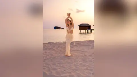 Elie stands on a sandy beach at sunset, wearing a white string bikini and raising her hands above her head.