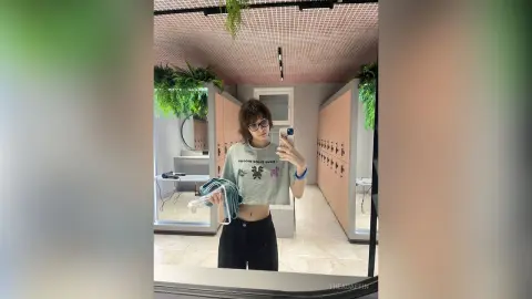 Young woman with glasses in a casual grey graphic tee taking a mirror selfie in a modern minimalist room.