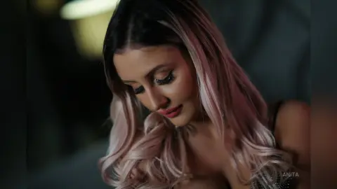 Anita looks down with long eyelashes and wavy blonde-highlighted hair, wearing black lace lingerie.