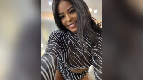 Smiling Black woman in a sparkly silver striped long-sleeve top posing indoors with a joyful mood.