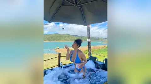 Woman in blue bikini relaxing in a bubble bath on a scenic lakeside terrace under a patio umbrella.