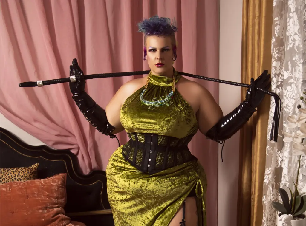 Smiling woman in a sparkling purple sequin gown and black harness posing against teal velvet curtains.