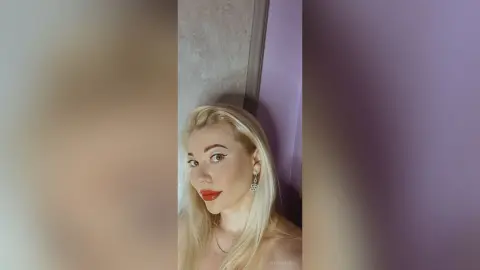 Blonde woman with winged eyeliner and red lipstick posing in a bright indoor setting.