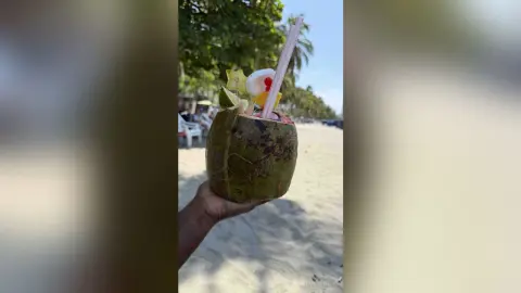 Hand holding a refreshing tropical coconut cocktail with fruit garnish on a sunny summer beach.