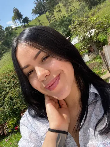 Smiling young woman with long black hair in a patterned shirt enjoying a sunny day in a lush green mountain field.