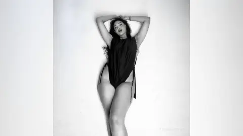 LaurenKaterine poses against a white wall wearing a black bodysuit, with her hands behind her head and her long dark hair down.