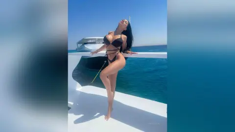 NicoleBella poses naked or in a black string bikini on a white yacht deck overlooking blue ocean water.