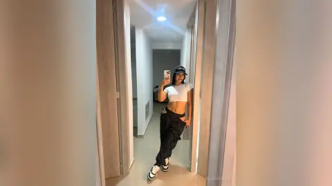 Young woman in white crop top and black cargo pants taking a mirror selfie in a modern hallway.