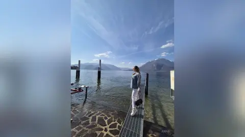 Woman in a blue sweater and white pants standing on a pier by a scenic lake under a bright blue sky.