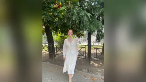 Elegant woman in a white flowing midi dress standing under lush green trees in a sunny outdoor park.