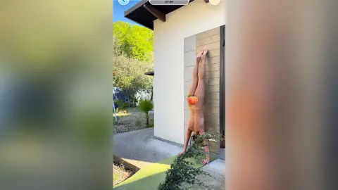 Fit woman in orange bikini performing a handstand outdoors near a modern house on a sunny day.