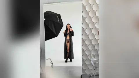 Fashionable woman in black lingerie and long coat posing in a bright professional photo studio.