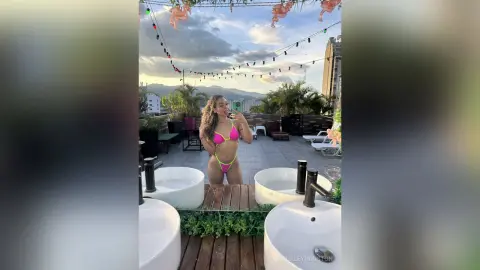 Young woman in a neon pink bikini taking a selfie on a sunny rooftop terrace overlooking a mountain landscape.