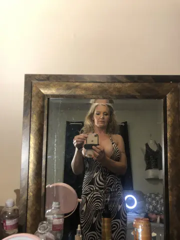 Blonde woman in a black and white swirl pattern jumpsuit taking a mirror selfie in a cozy bedroom setting.