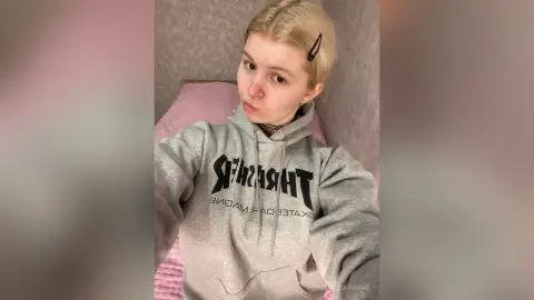 Blonde woman in a grey Thrasher hoodie posing in a cozy bedroom setting.