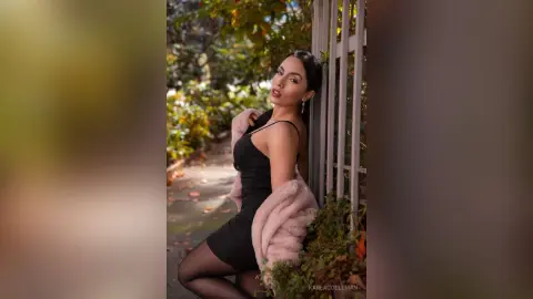 Elegant woman in a black dress and pink faux fur coat posing outdoors in a garden setting.