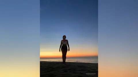 Silhouette of a woman in a white crop top and black shorts standing on a beach during a golden sunset.
