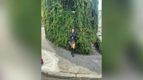 Young woman in a black leather jacket and mini skirt standing by a lush green ivy wall on a city street.