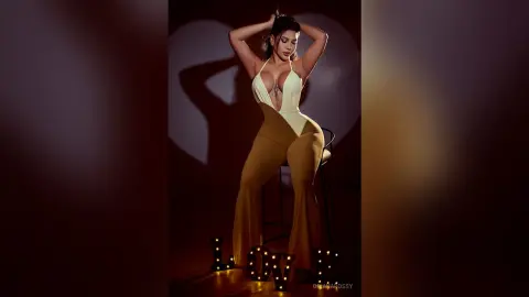Glamorous woman in a white halter bodysuit and tan flared trousers posing against a romantic heart shadow.