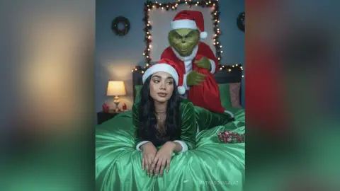 Young woman in green velvet outfit relaxing on a bed next to the Grinch in a festive holiday setting.