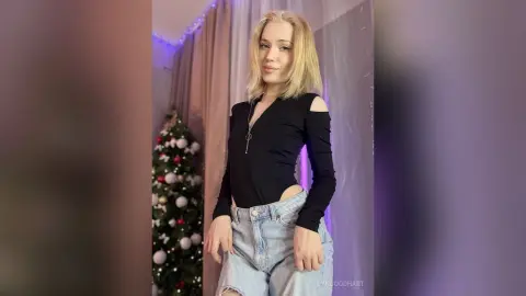 Blonde woman in a black long-sleeve bodysuit and light denim jeans posing by a decorated Christmas tree.