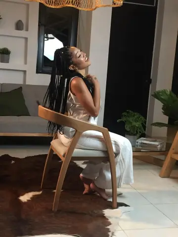 Joyful Black woman in white loungewear sitting on a wooden chair in a modern, sunlit living room.