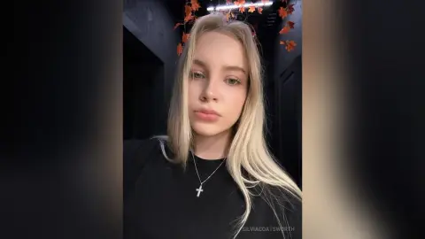 Blonde woman in a black t-shirt and silver cross necklace posing with a moody, soft-lit aesthetic.