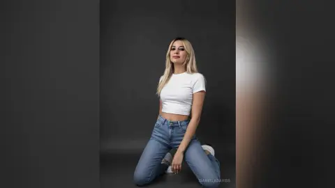 Blonde woman in a white crop top and blue jeans posing confidently against a dark studio background.