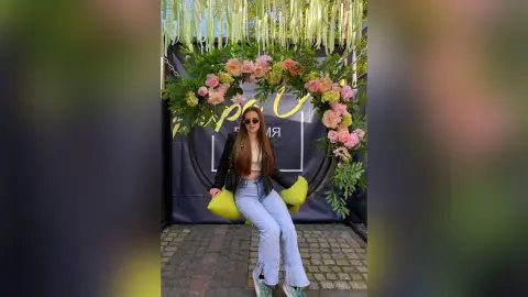 Young woman in sunglasses and jeans sitting on a floral swing decorated with pink roses in a garden setting.