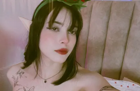 Young woman with elf ears and dark hair posing in a cozy bedroom setting with a soft, dreamy mood.