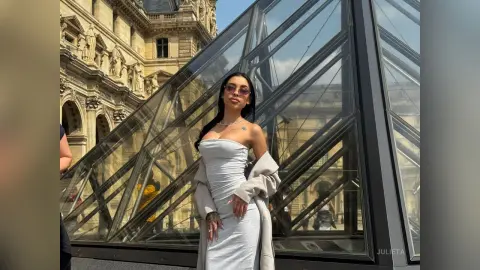 Stylish woman in a grey strapless dress posing by the Louvre glass pyramid in Paris on a sunny day.