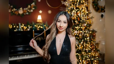 Smiling woman in a black sequin dress posing by a glowing Christmas tree in a cozy festive setting.