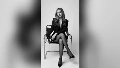 MeganLewit sits on a chair wearing a black blazer and sheer black tights with high heels.