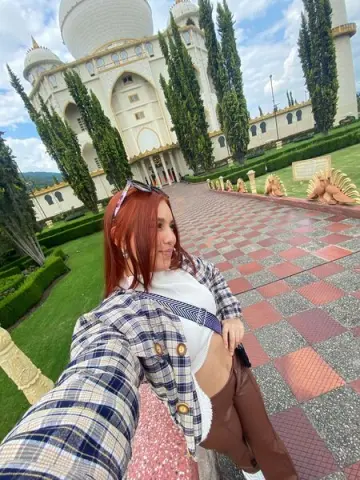 Red-haired woman in a plaid blazer and white top posing at the Baha'i Temple courtyard under a blue sky.