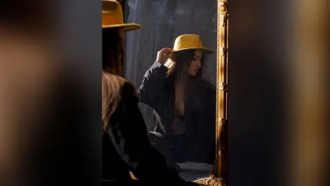 Moody portrait of a woman in a dark blazer and yellow wide-brimmed hat reflected in a vintage gold mirror.